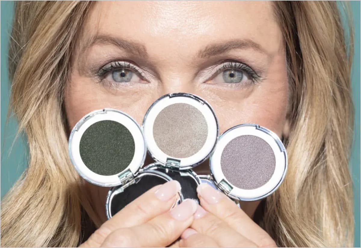 Blonde woman with blue eyes holds three open compacts in dark green, beige and mauve shimmer over her face