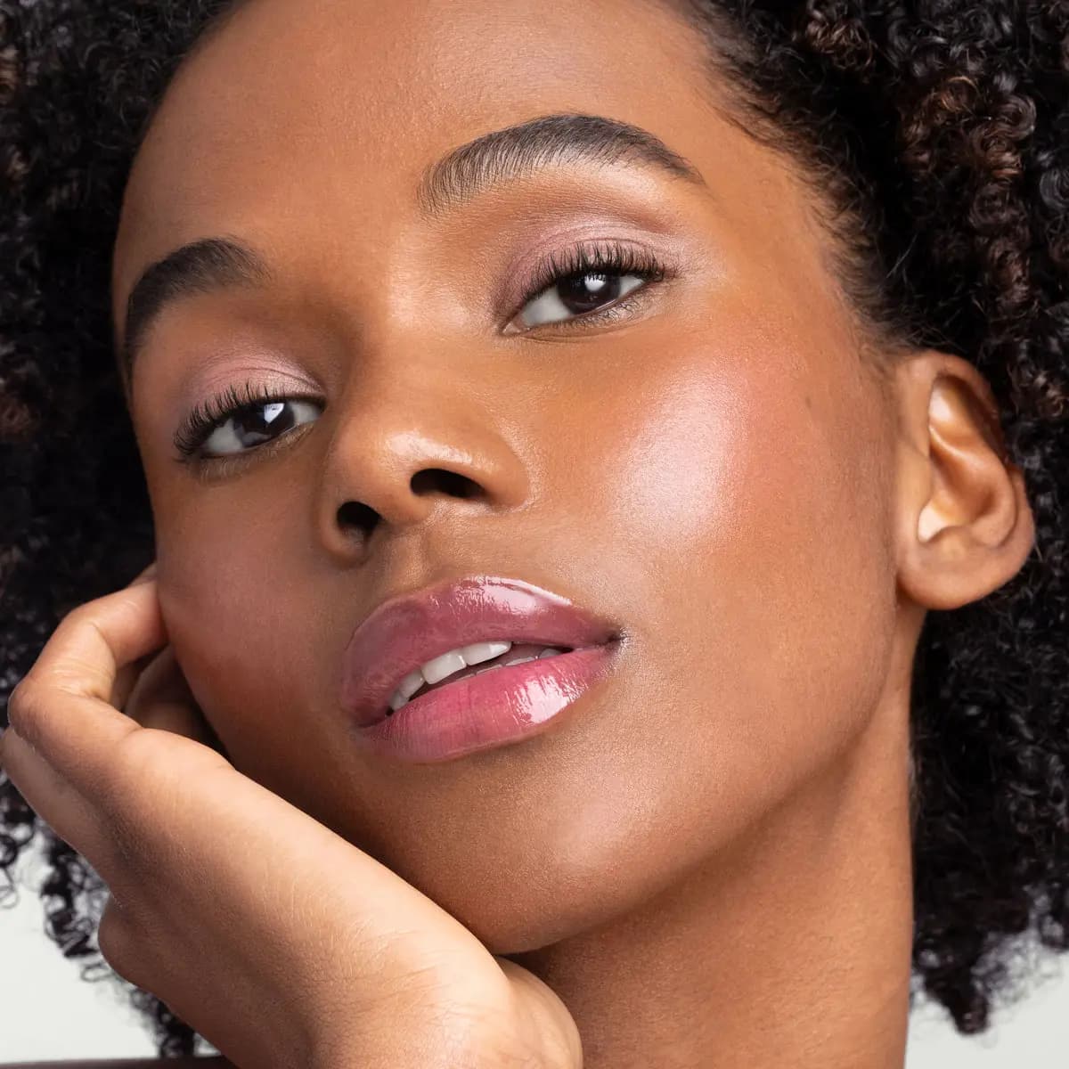 Model with curly dark hair rests her face on her hand, wearing glossy rosy-mauve lips and soft pink eyeshadow.