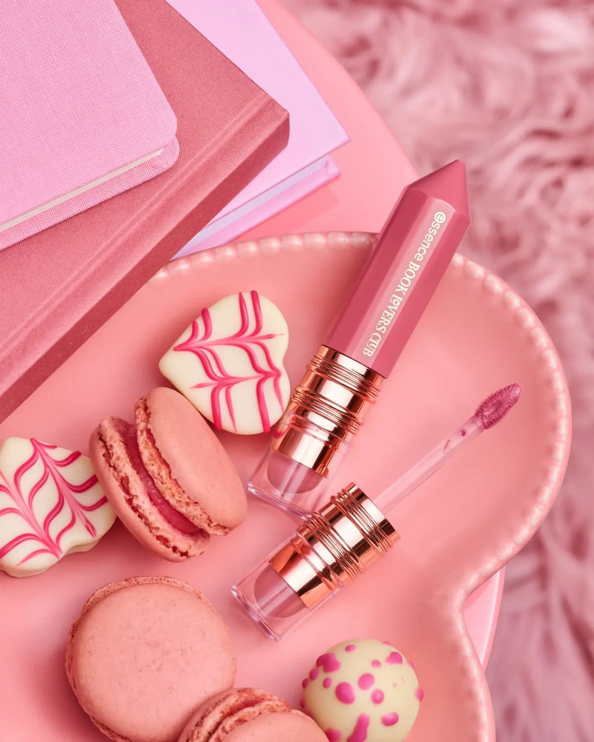 Pink lip cream with crayon-shaped cap and doe-foot wand on a plate with macarons and heart chocolates