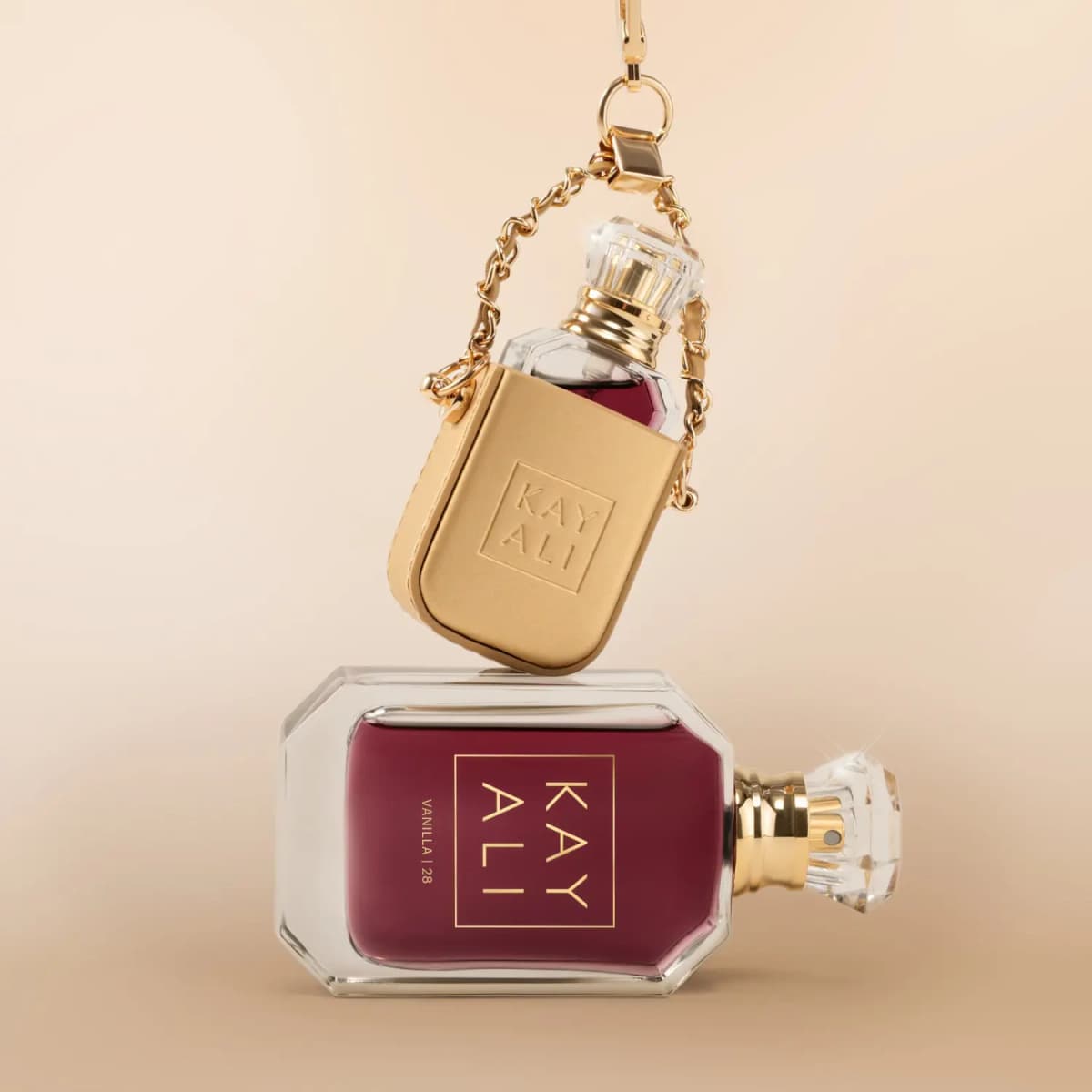 Gold holder with embossed Kayali logo hanging on a chain above the burgundy Vanilla | 28 bottle on a beige backdrop