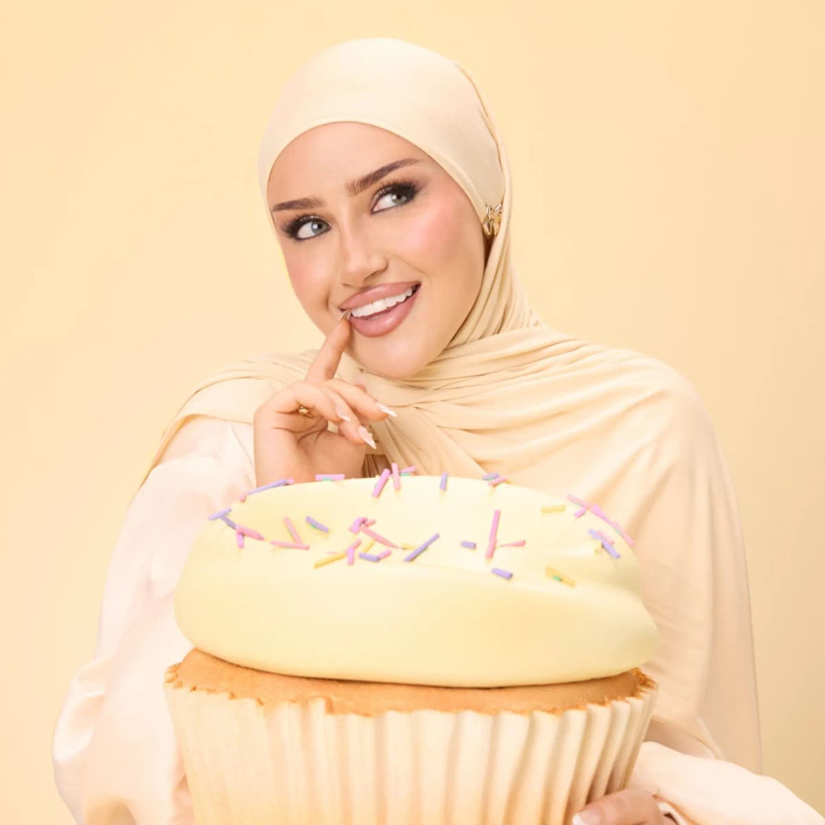 Model in a cream hijab smiles playfully while hugging an oversized vanilla cupcake topped with pastel sprinkles
