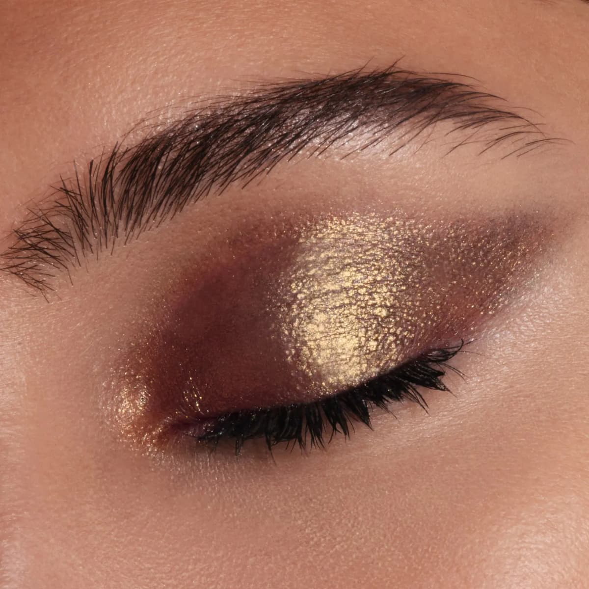 Closed eye with a deep burgundy-brown wash and a bright gold metallic shimmer patch on the centre of the lid