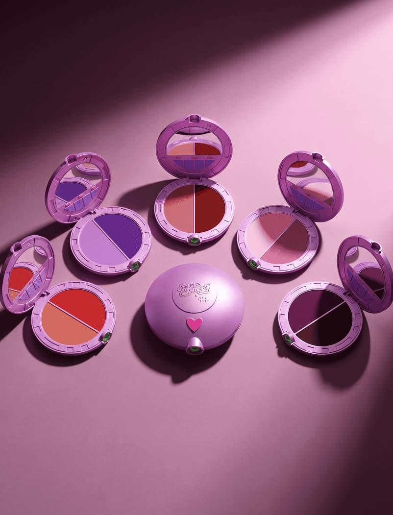 Seven pink round compact blush duos arranged in a circle with a closed heart-embossed compact centered on pink surface.