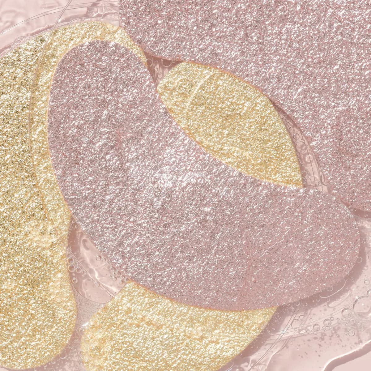 Overlapping shimmering gold and rose gold foil eye masks resting in glossy clear serum with tiny bubbles