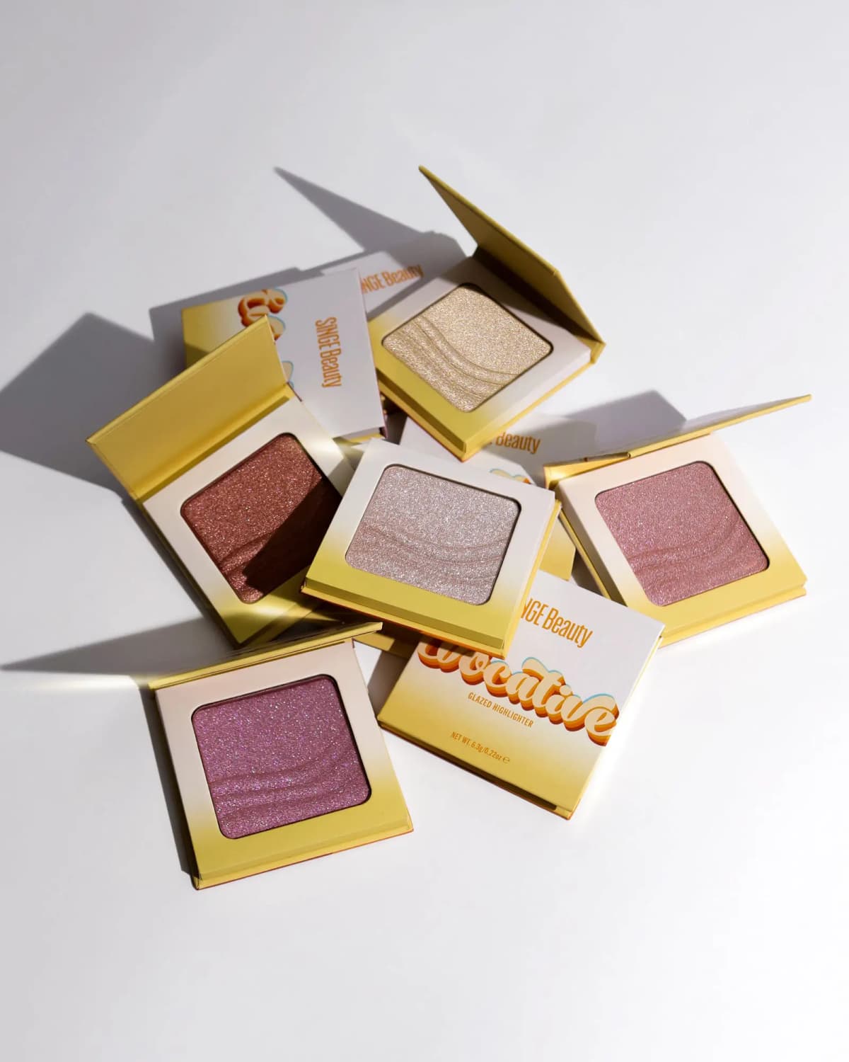 Five open glazed highlighter compacts scattered with long shadows on white surface