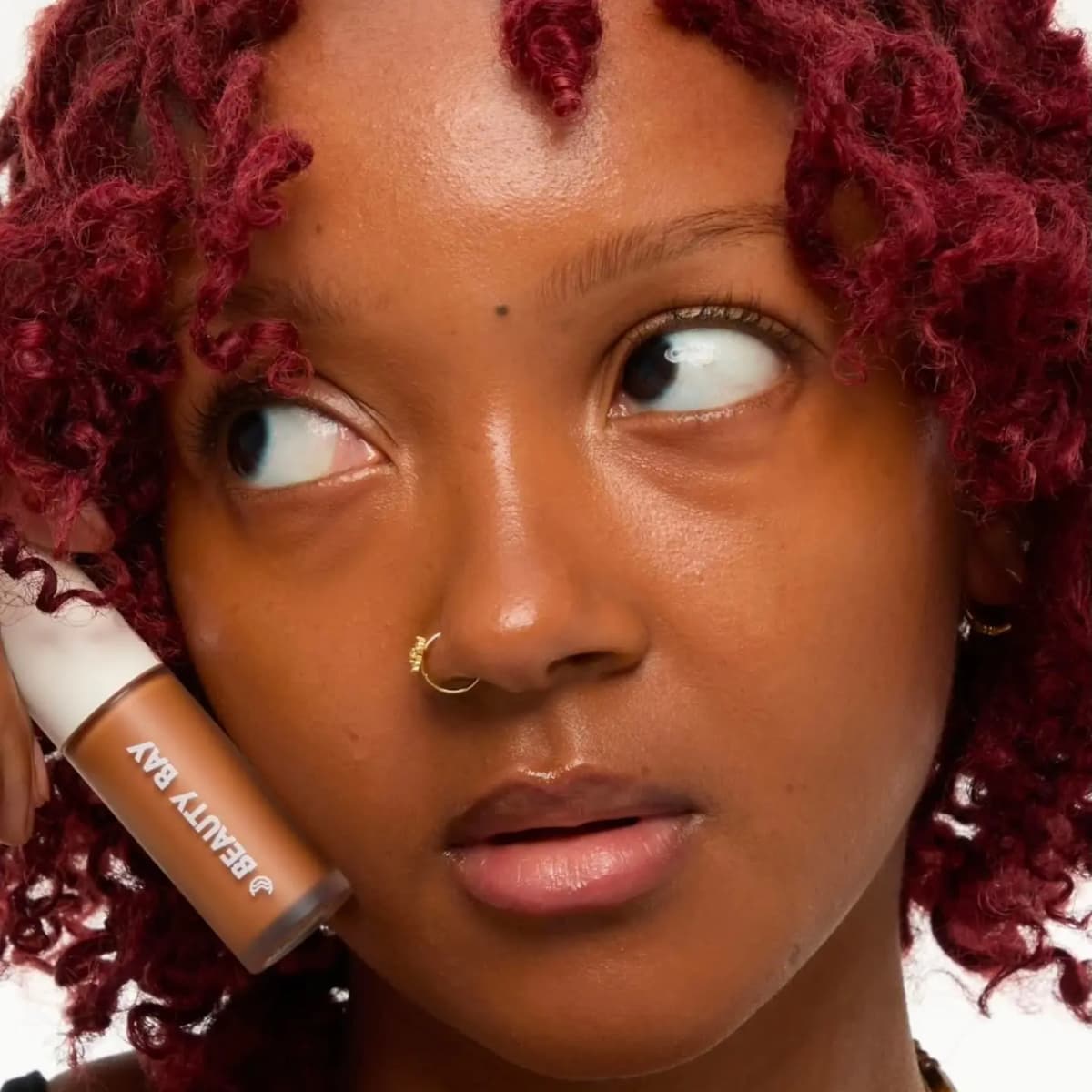 Model with deep skin and red curly locs holds a brown Beauty Bay concealer bottle against her cheek