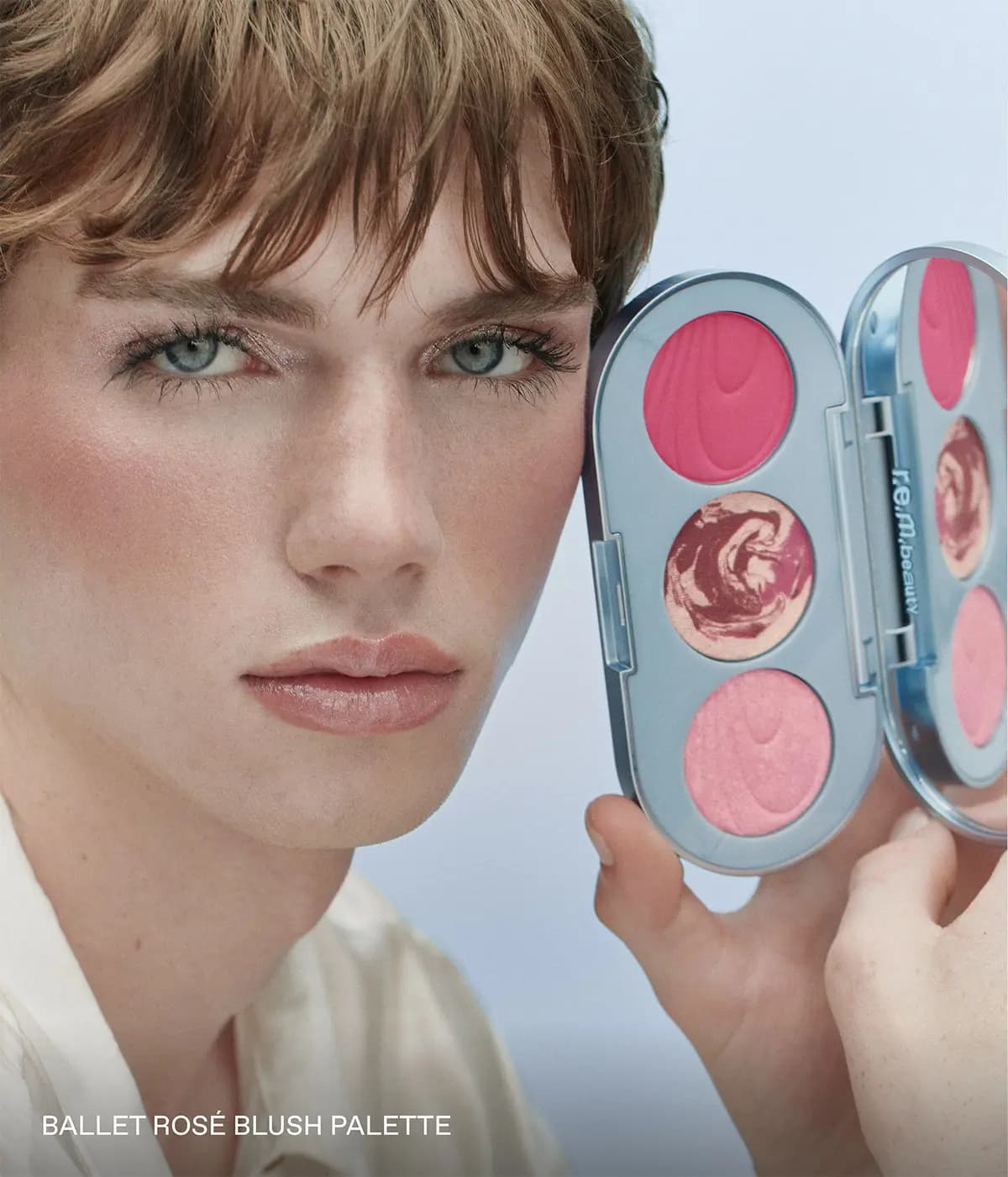 Male model with rosy flushed cheeks and glossy lips holds an open Ballet Rosé blush palette beside his face