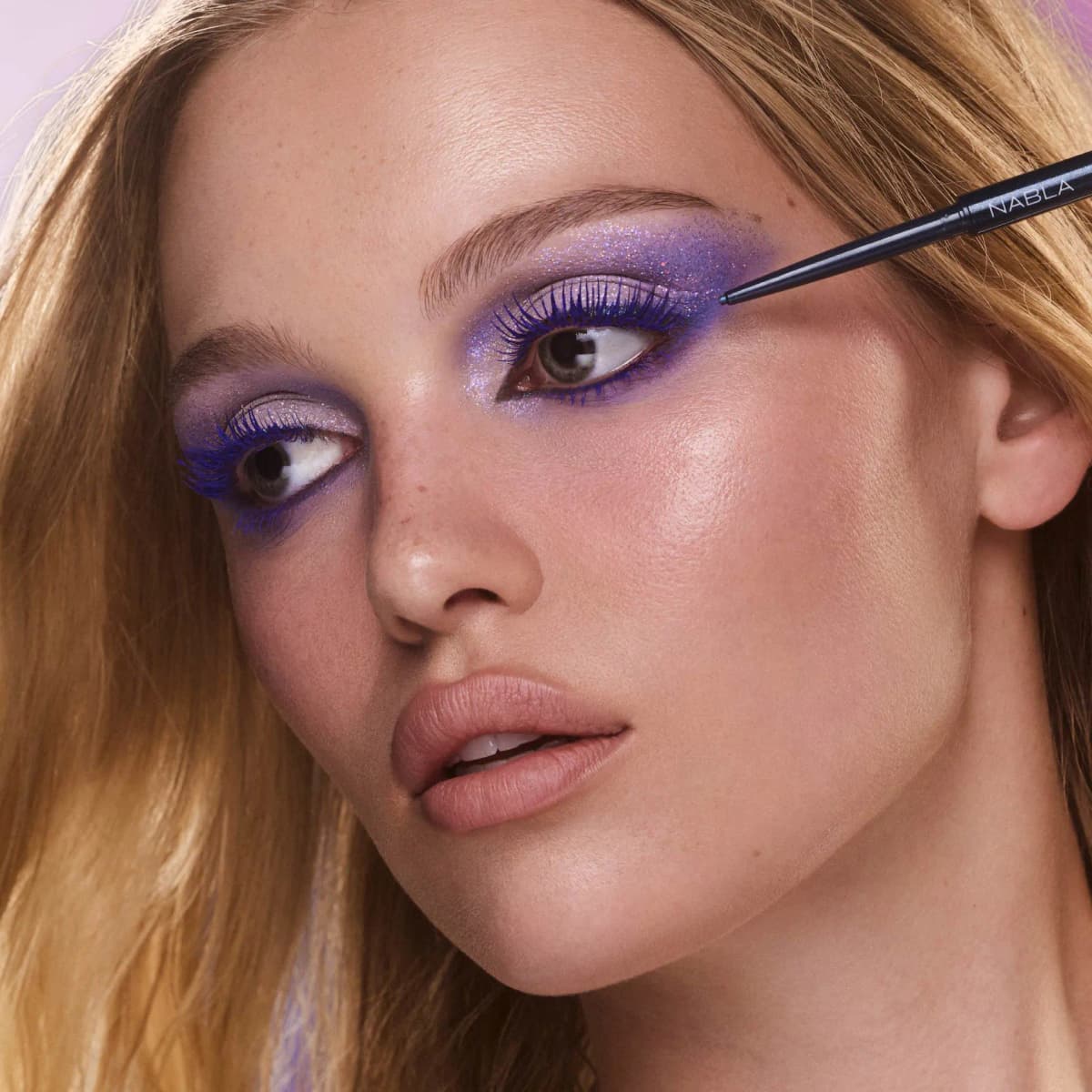 Model with violet glitter shadow and blue lashes lines her outer eye with the navy NABLA pencil.