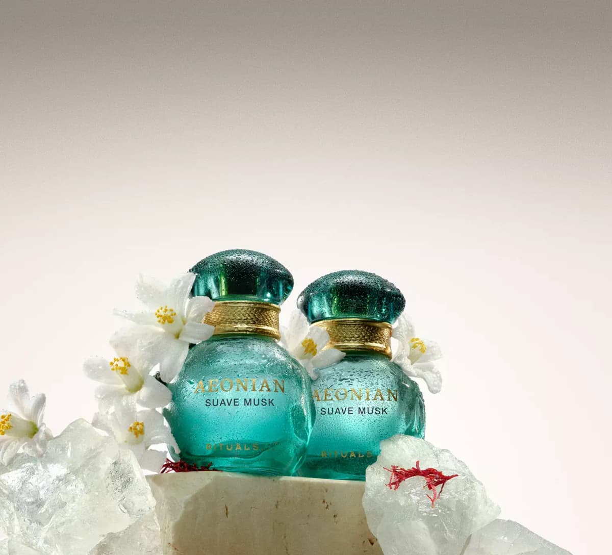 Two dewy aqua Rituals Aeonian Suave Musk bottles with white blossoms, clear crystals and saffron threads on stone