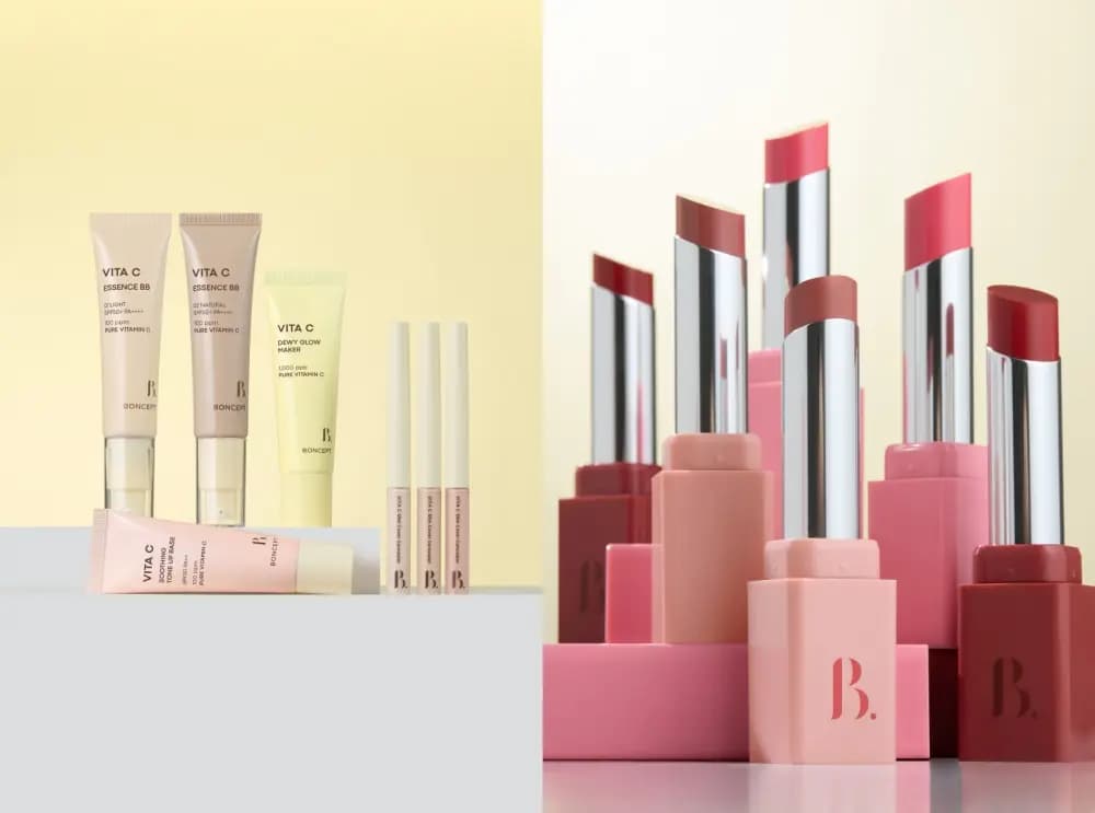 BONCEPT Vita C makeup line base tubes and concealers on a yellow backdrop beside pink and red lipsticks on pink blocks