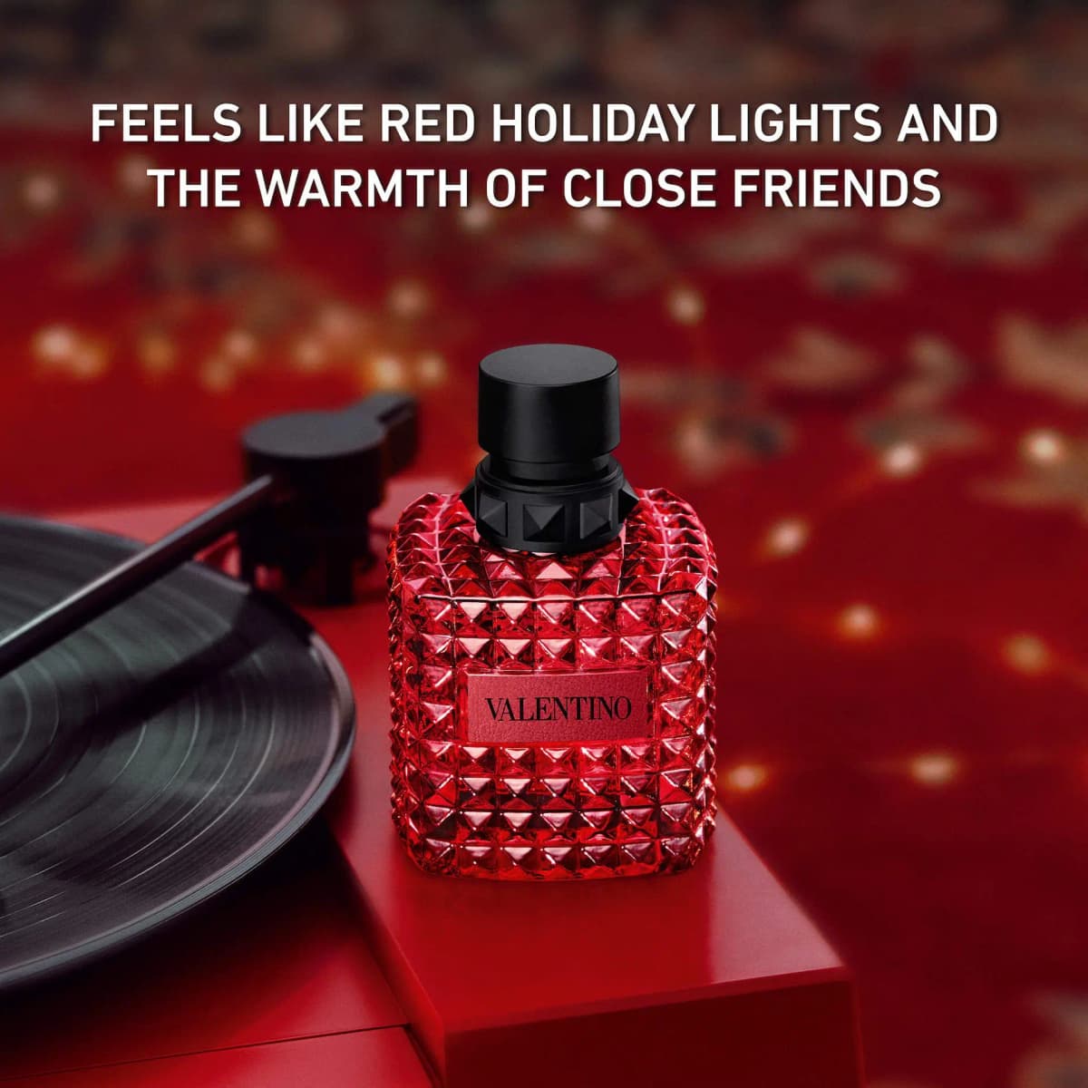 Red faceted Valentino Born in Roma bottle beside a vinyl record on a red surface with bokeh lights.
