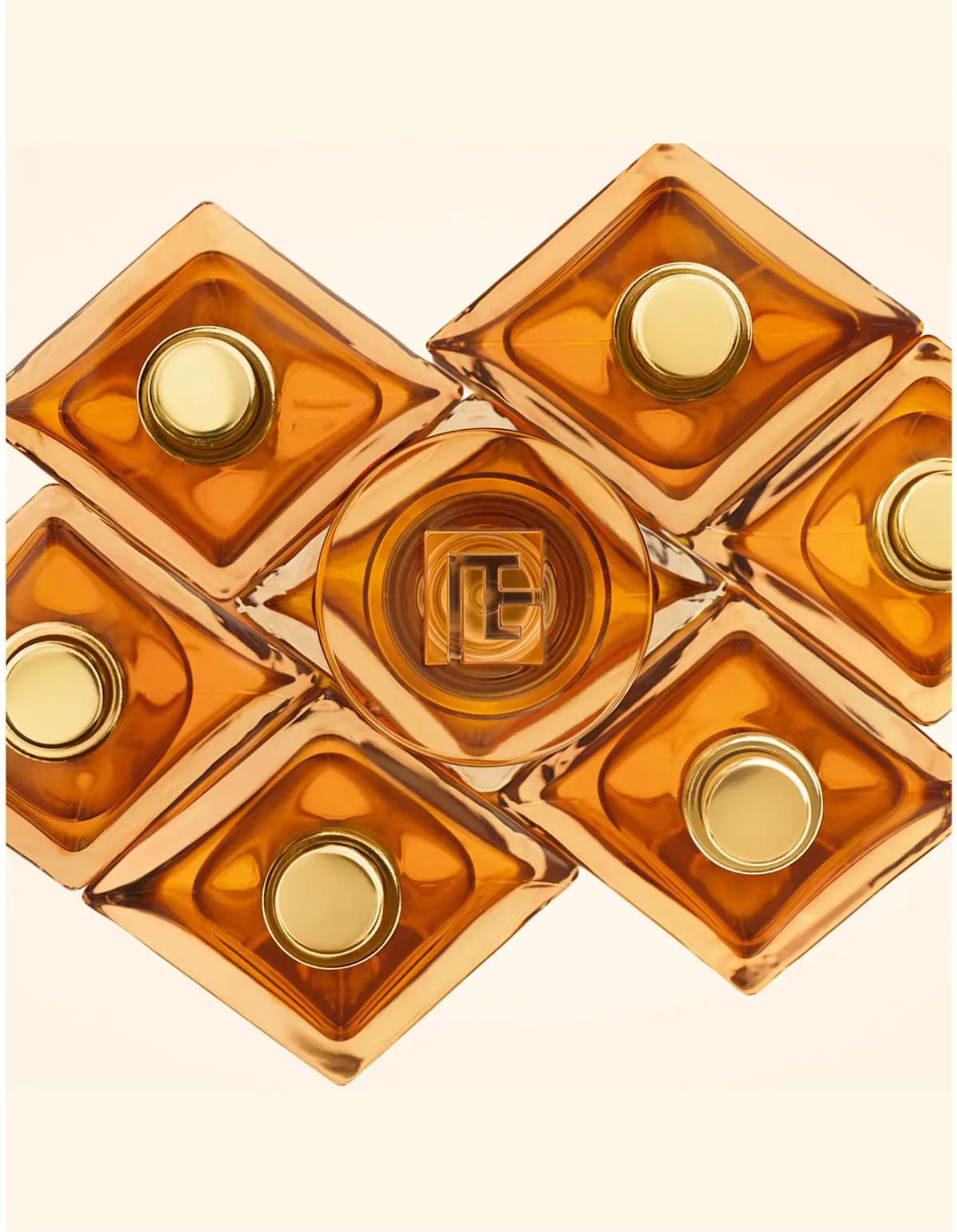 Overhead view of amber bottles with gold caps arranged in a diamond pattern around a cap bearing the Balmain monogram