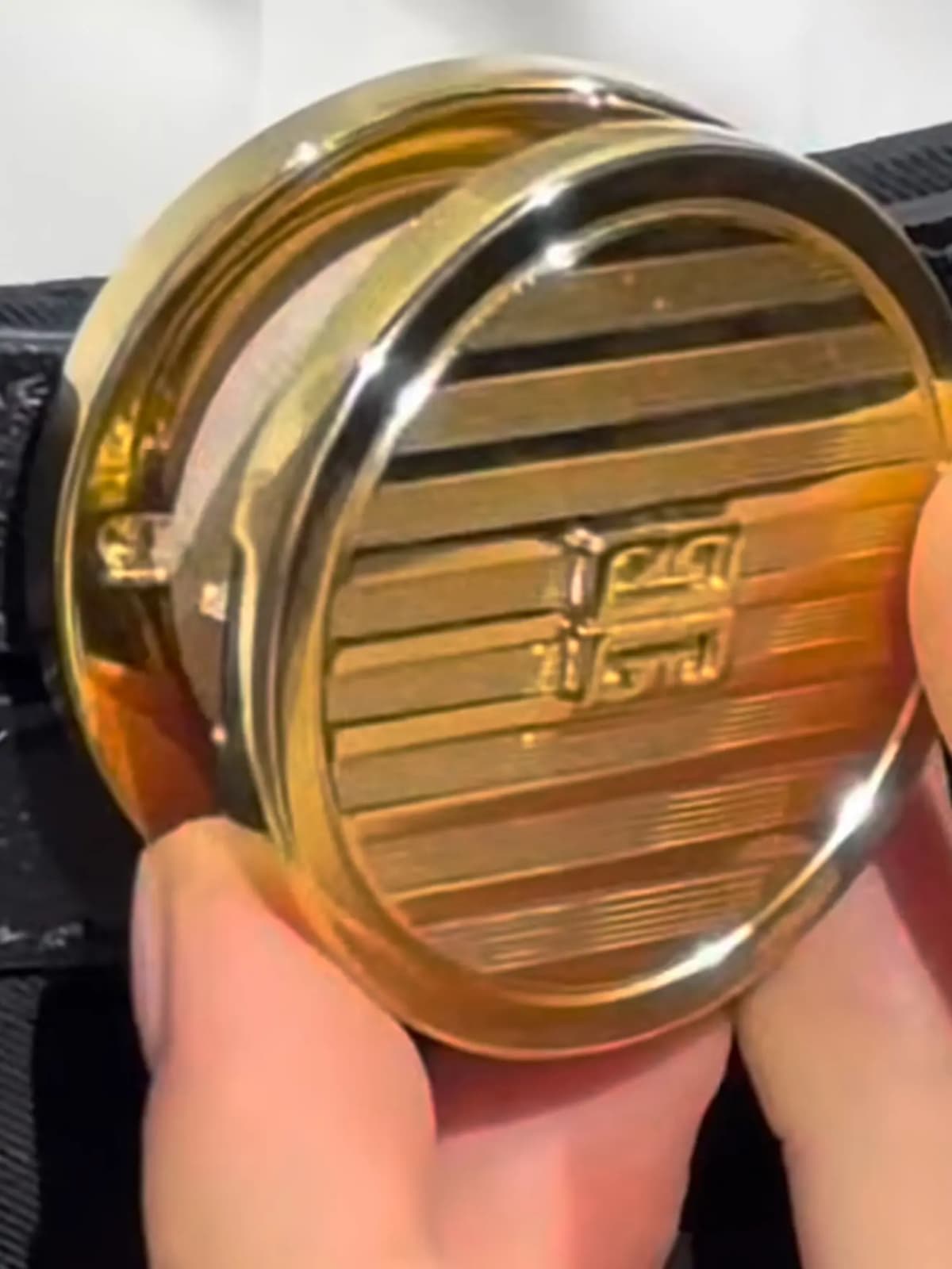 Close-up of a hand holding a gold compact with a striped engraved lid and raised 4G logo
