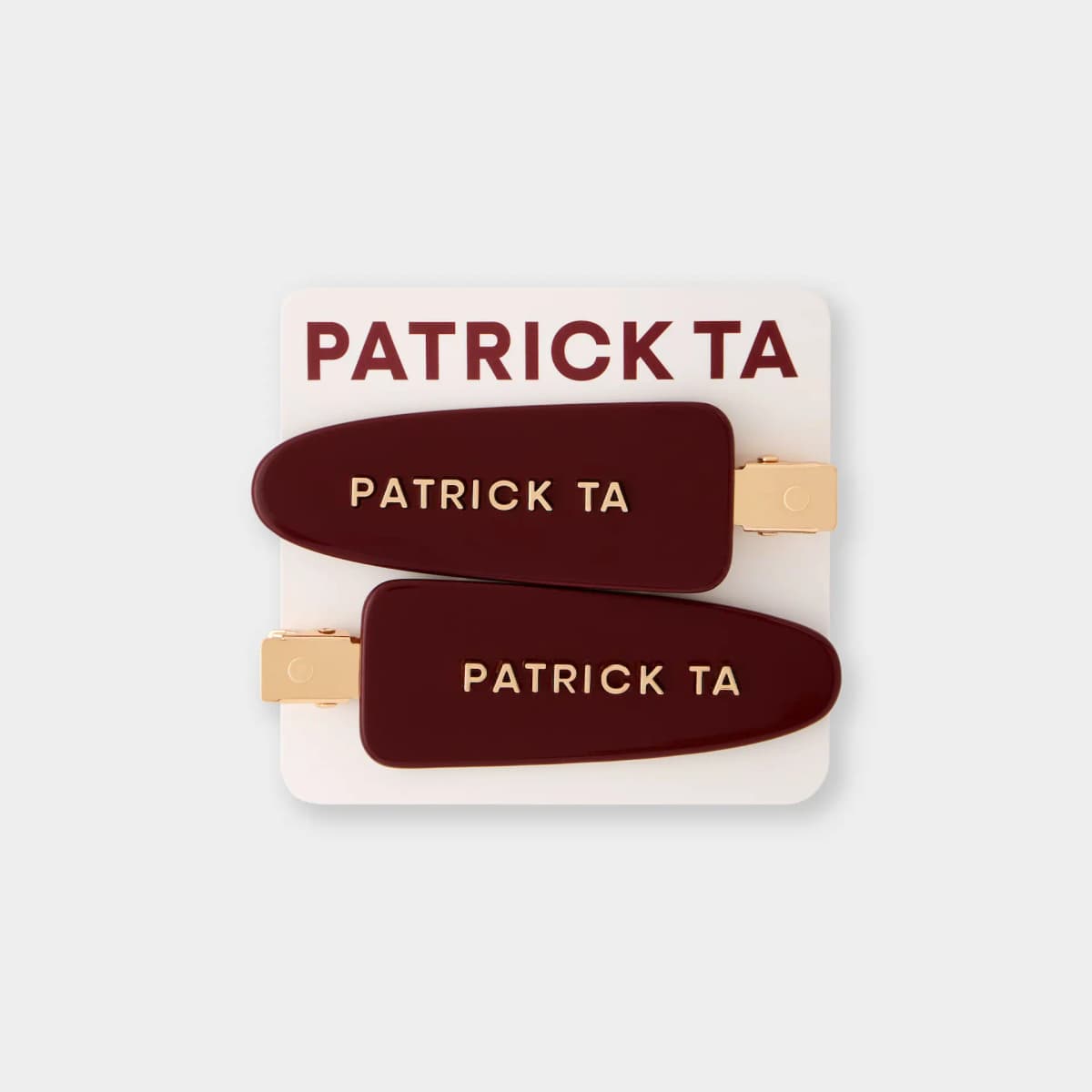 Two glossy burgundy hair clips with gold Patrick Ta lettering and gold clasps on a cream branded card