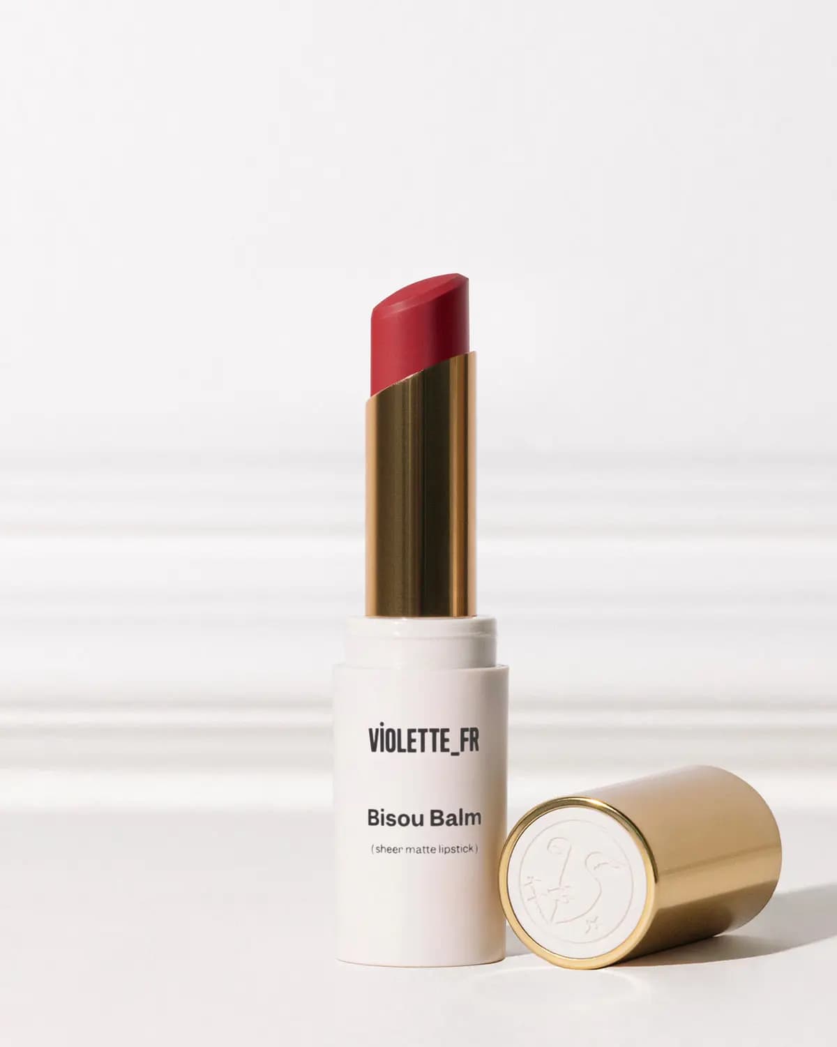 Violette_FR Bisou Balm sheer matte lipstick in a red-rose shade, white tube with gold cap lying beside it.