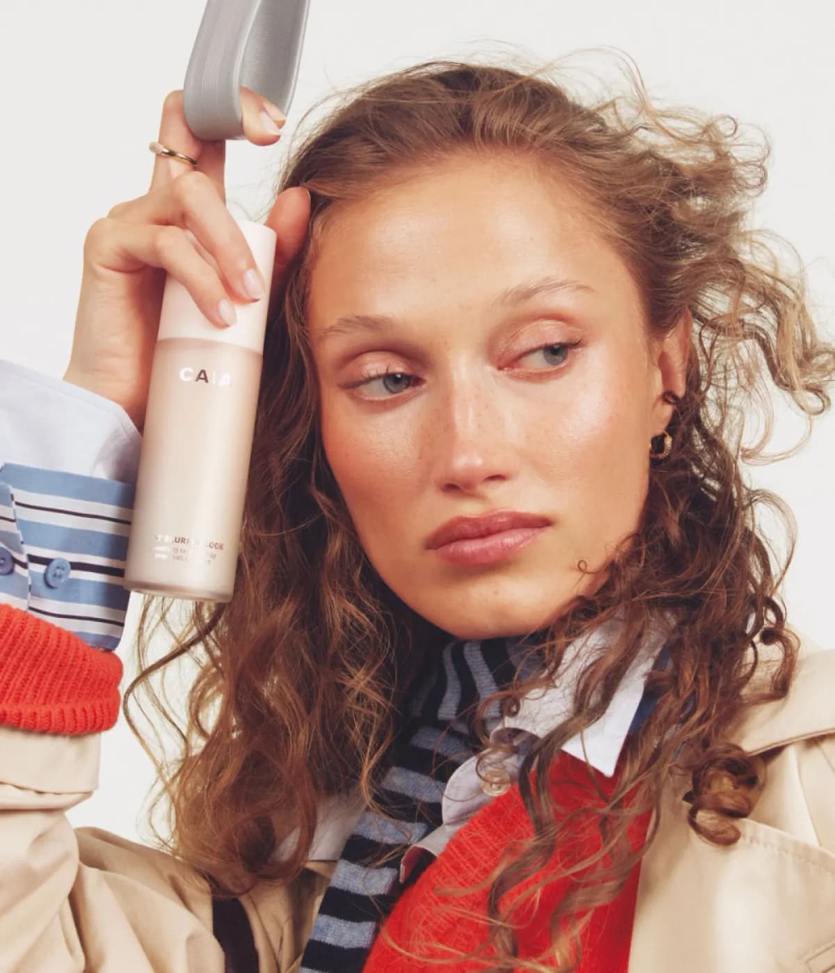 Curly-haired freckled woman in a trench coat and red knit holds CAIA That Blurred Look near her temple