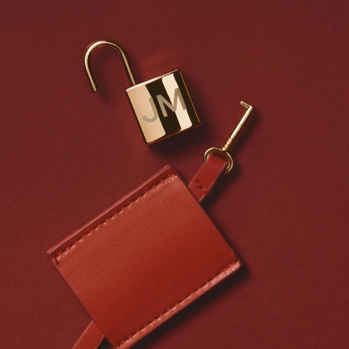 Gold padlock engraved JM lying open beside its small key on a red leather tag against a deep red backdrop
