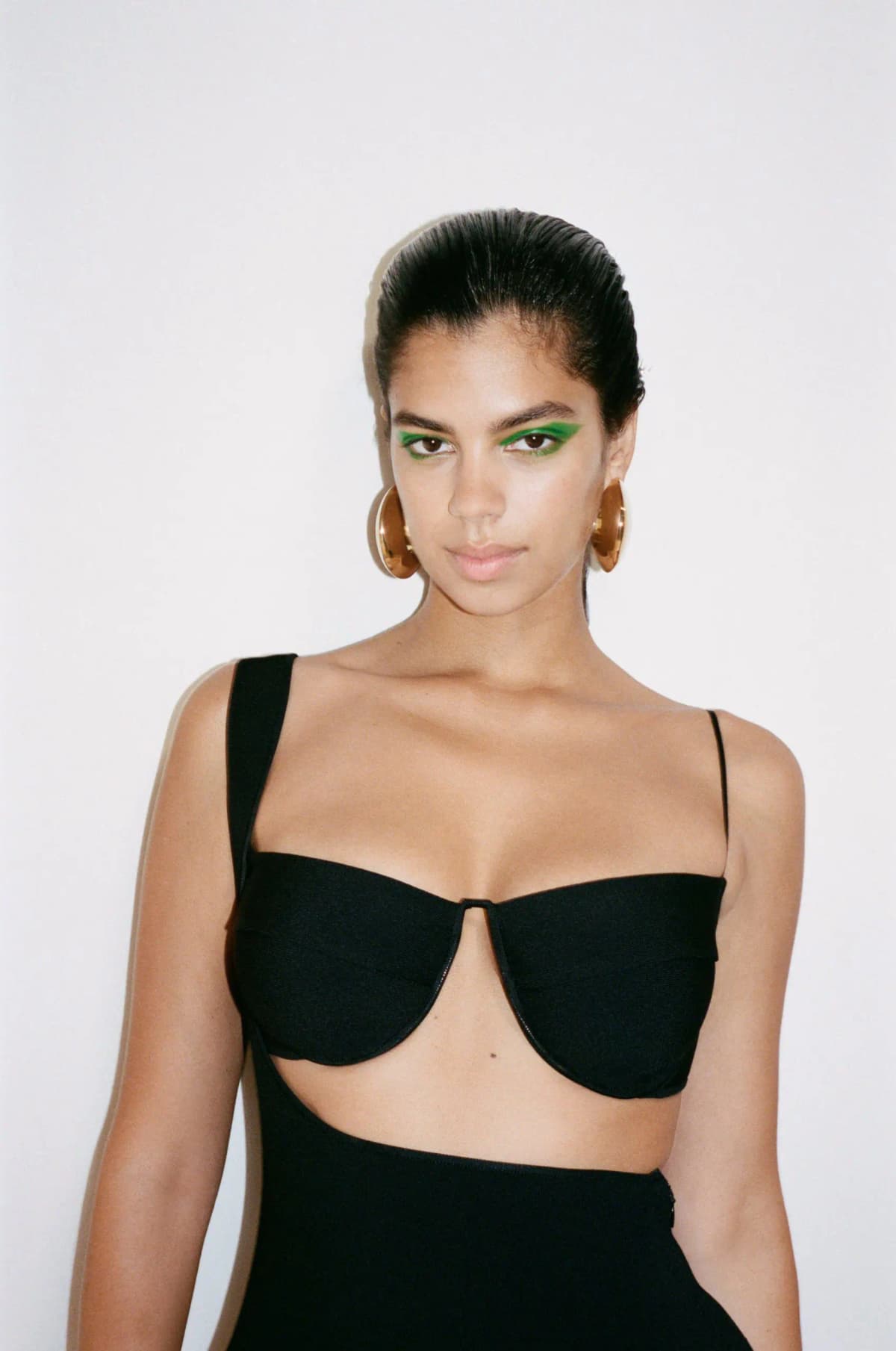 Model with bold green eyeliner, slicked-back hair, gold disc hoops and a black cutout underwire top