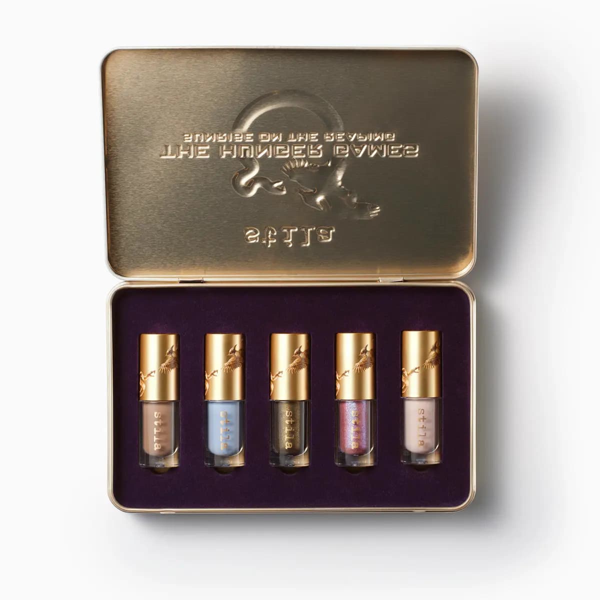Open gold tin with embossed lid holding five mini shadow vials on purple velvet, from taupe and blue to shimmery pink