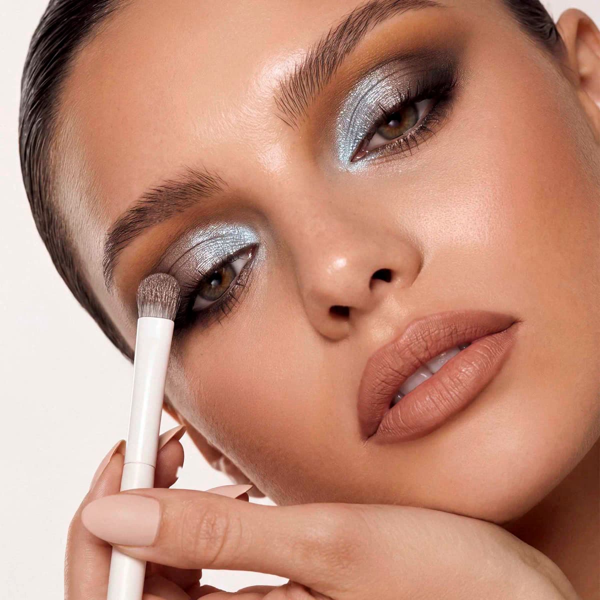 Close-up of a model applying a smoky look of icy blue shimmer and dark brown with a white eyeshadow brush.