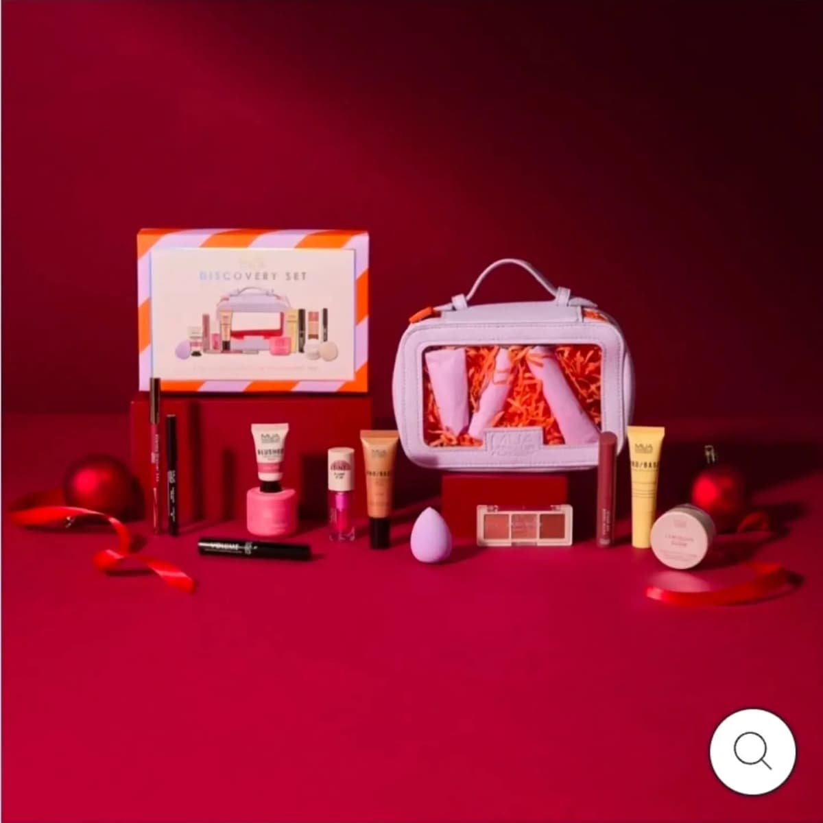 MUA Discovery Set box beside a lilac clear-window makeup bag, surrounded by minis, a purple sponge and red baubles.