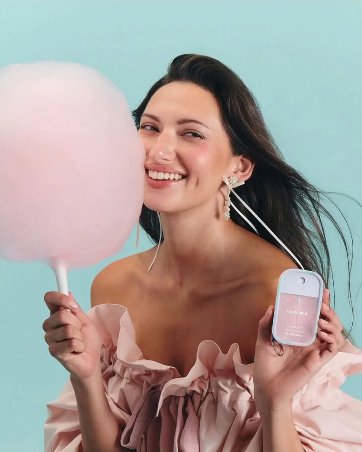 Smiling brunette woman in ruffled pink top holding pink cotton candy and a pastel Touchland Power Mist against teal backdrop