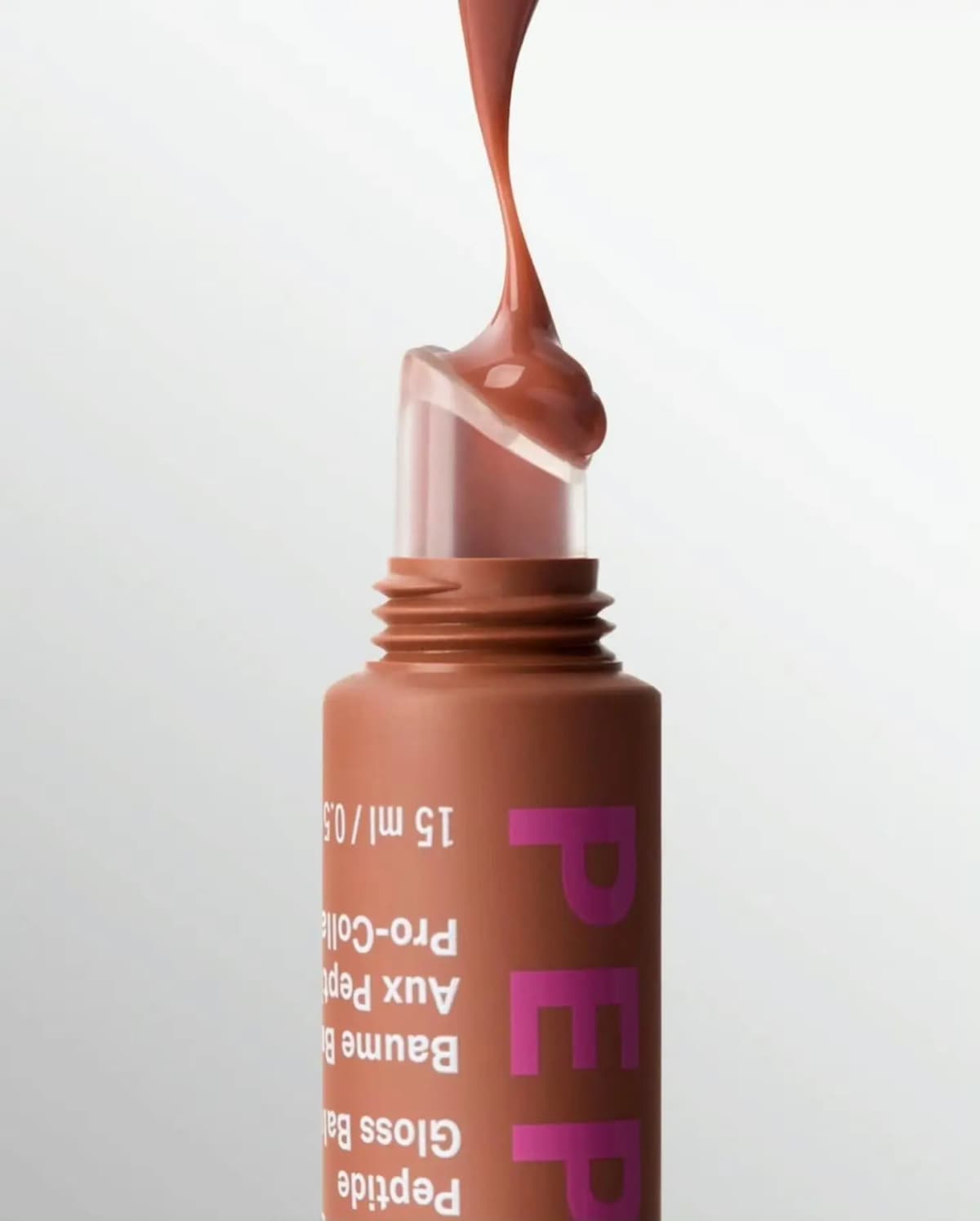 Glossy warm brown balm drips onto the clear slanted applicator tip of an upside-down brown tube