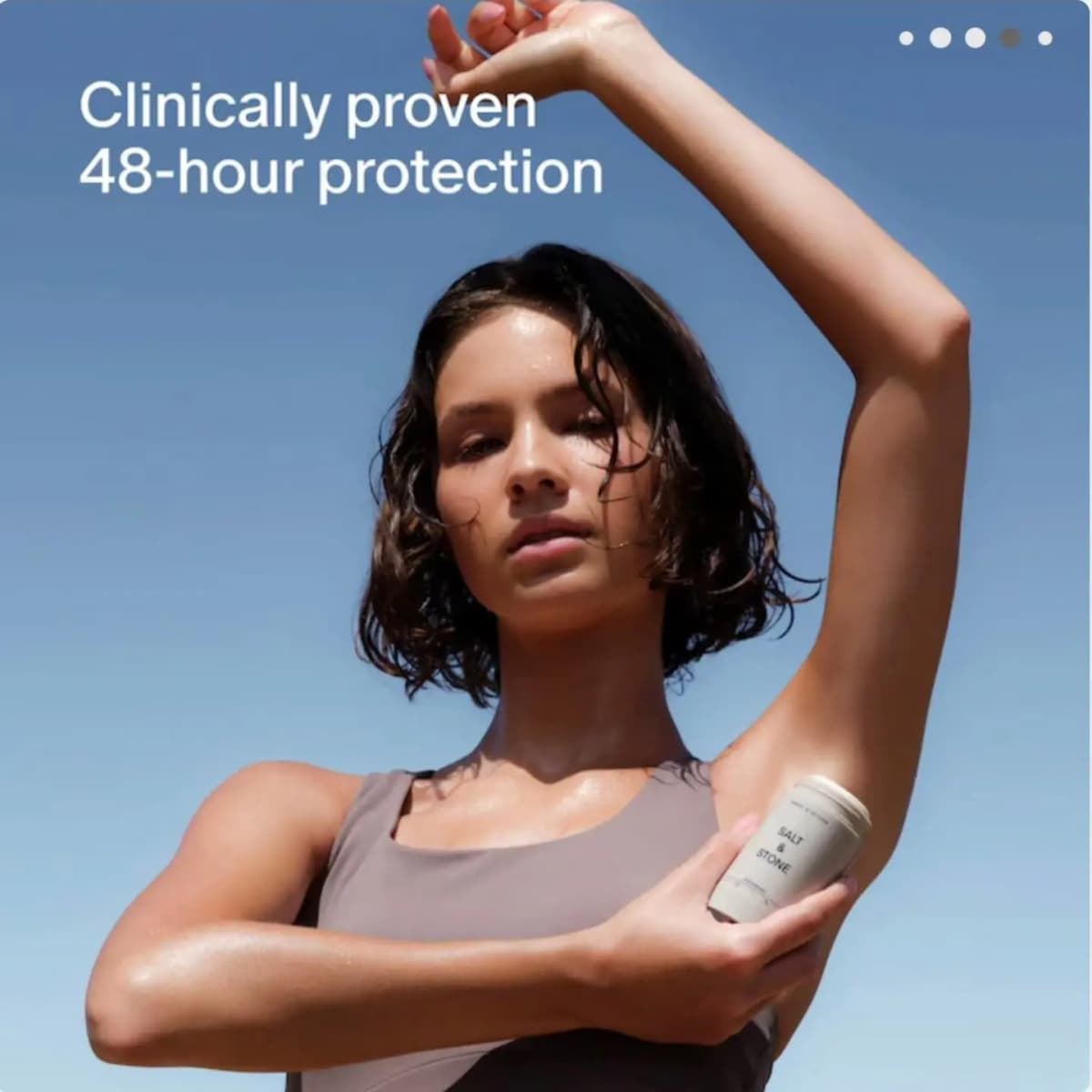 Woman with wet hair against blue sky raising one arm and holding a cream Salt & Stone deodorant under her underarm.