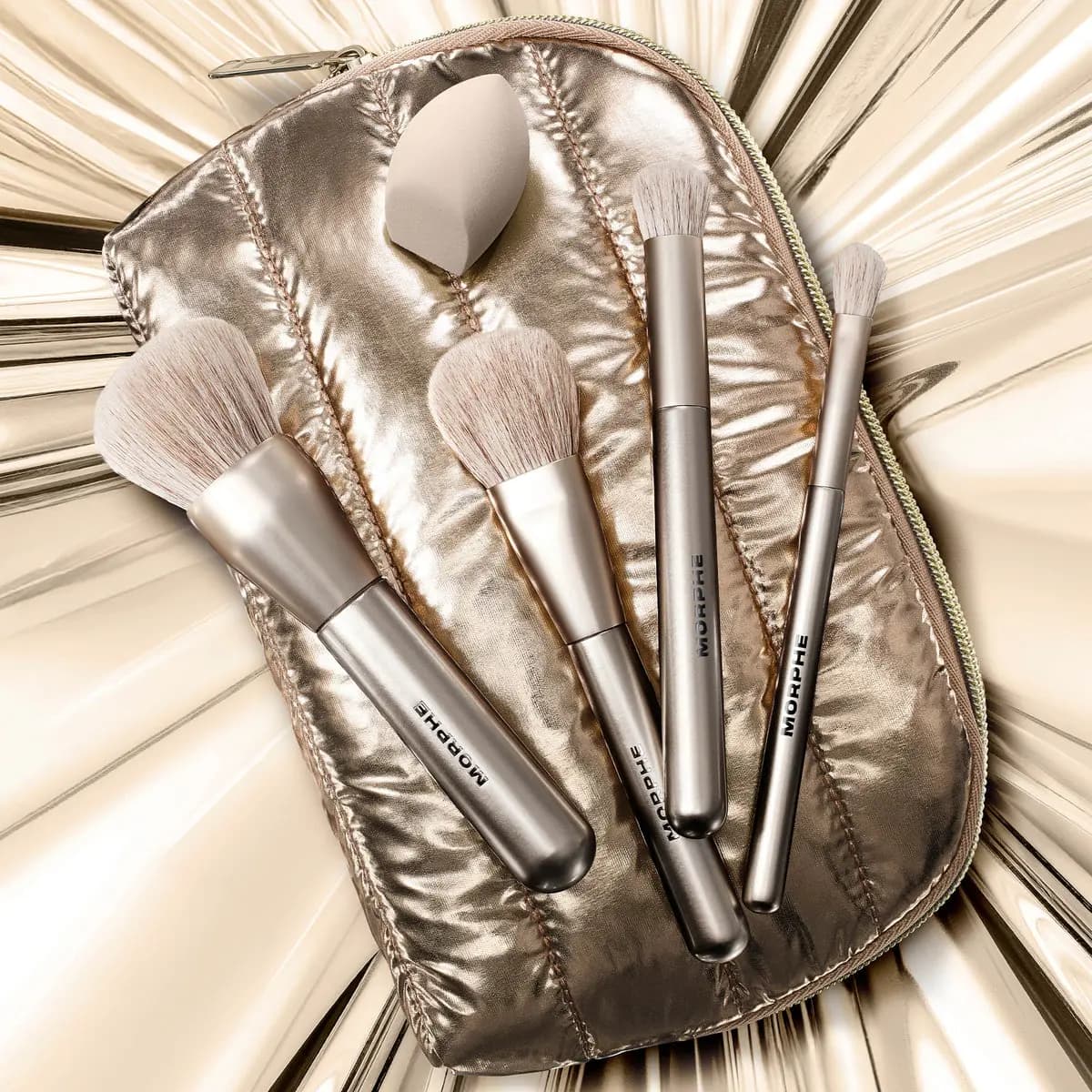 Four champagne Morphe brushes and a beige makeup sponge resting on a quilted metallic pouch over gold foil