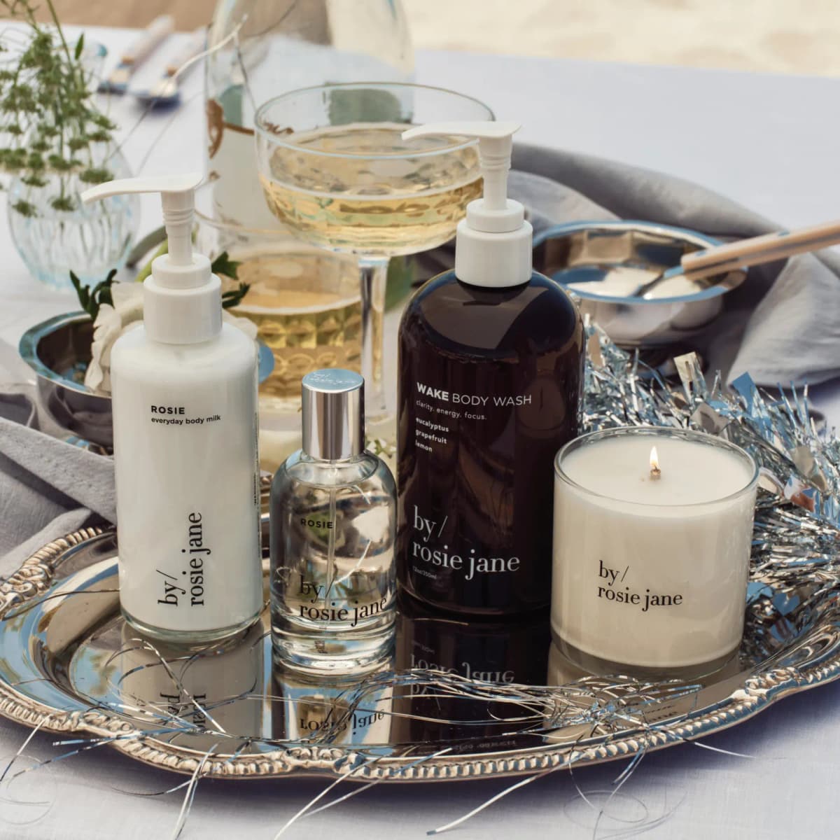 Rosie body milk, Rosie perfume, dark Wake body wash and lit candle on a silver tray with tinsel and a coupe glass