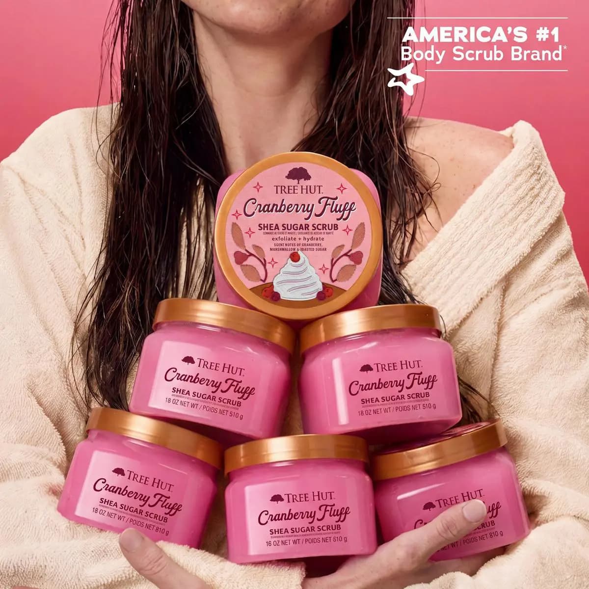 Woman in cream robe holding a pyramid stack of pink Cranberry Fluff Shea Sugar Scrub jars on pink background