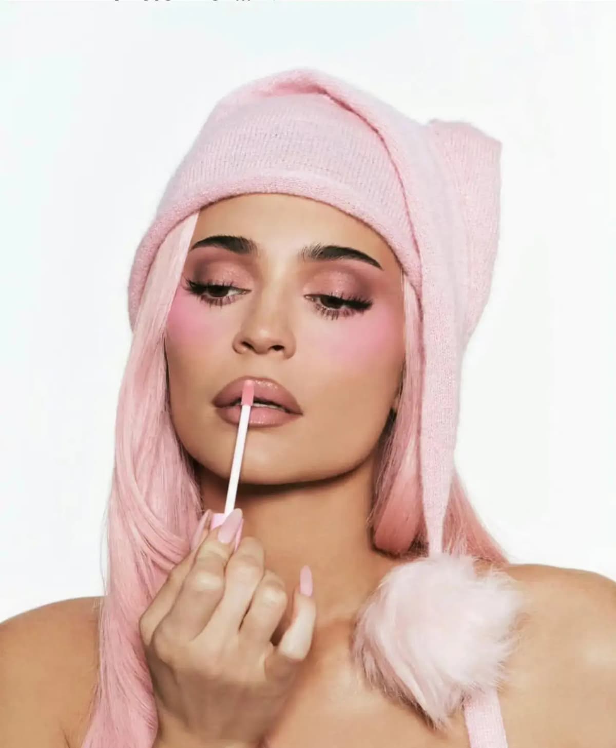 Kylie Jenner in a pink knit Santa hat and pink wig applies nude-pink lip gloss, with rosy blush and shimmery pink eyes