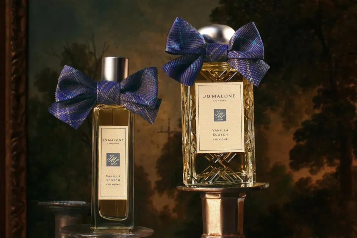 Jo Malone London Vanilla Scotch Cologne in two bottles with purple tartan bows, set against a dark landscape painting