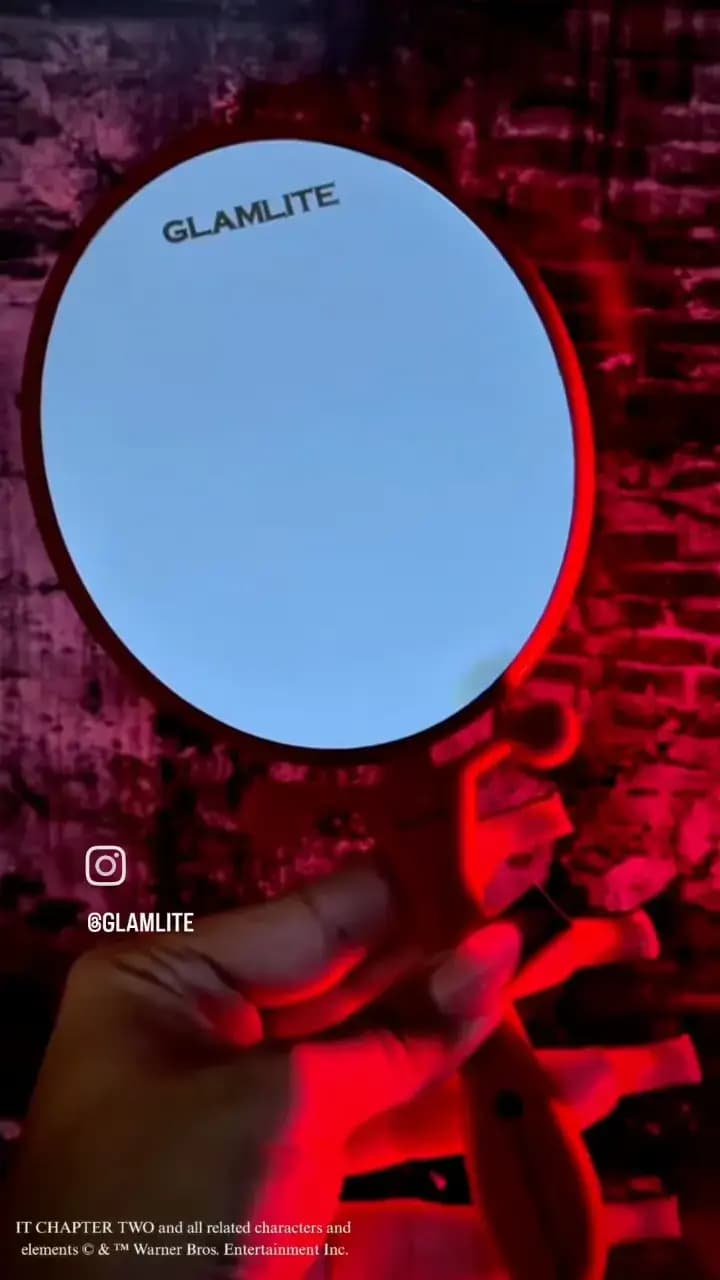 Hand holding the open red balloon-shaped mirror, its reflective side printed with Glamlite, under red light