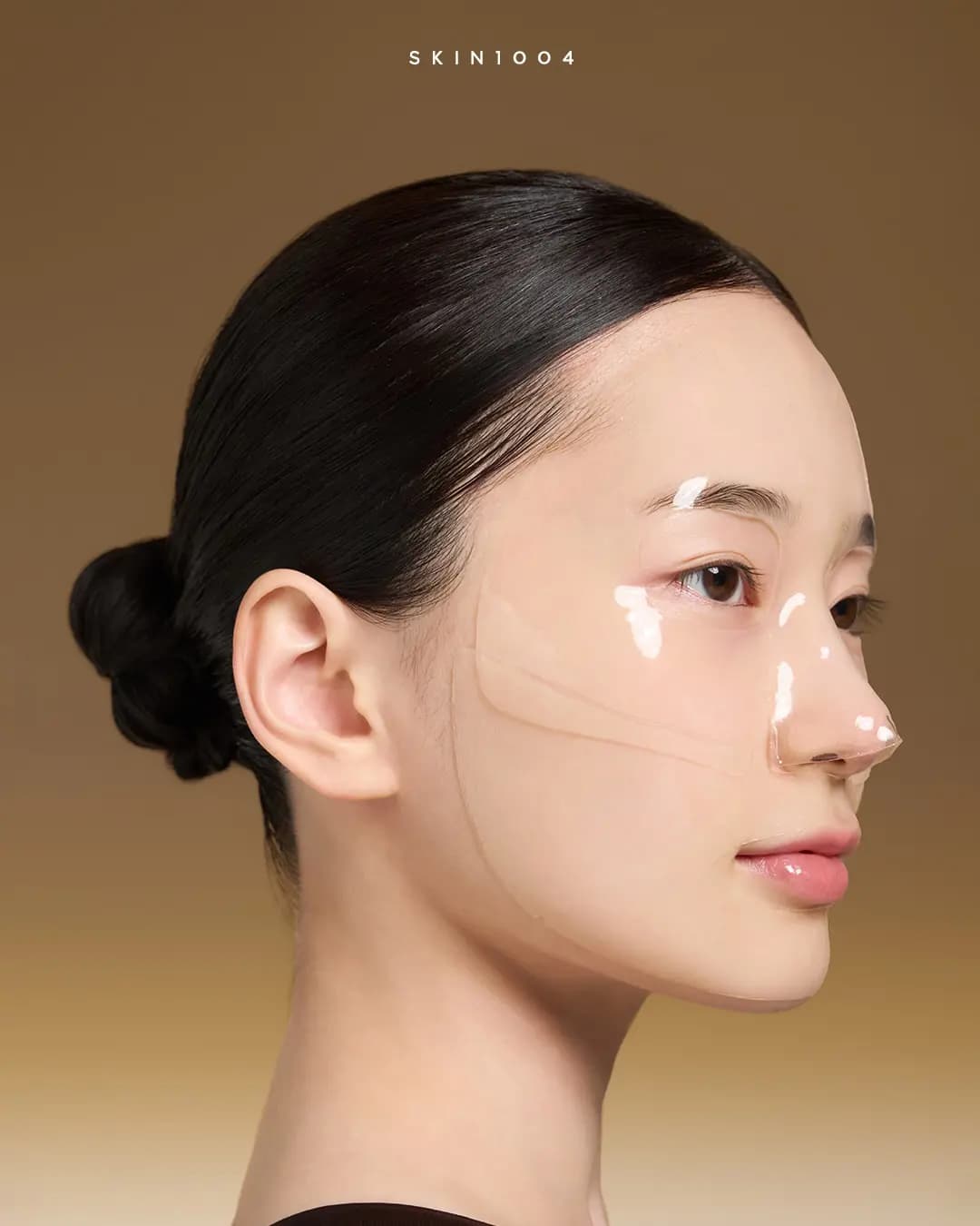 Model in profile wearing a clear glossy gel mask over the cheek, nose and jaw against a warm brown backdrop