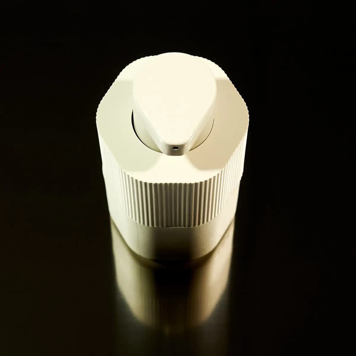 Top-down view of a cream Pump Capsule showing the teardrop-shaped actuator and nozzle hole on a dark surface
