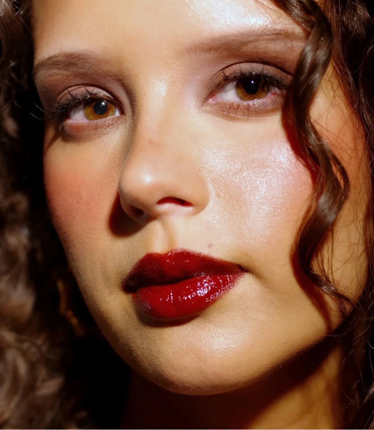 Close-up of a curly-haired model with amber eyes wearing a high-shine, juicy deep red lip