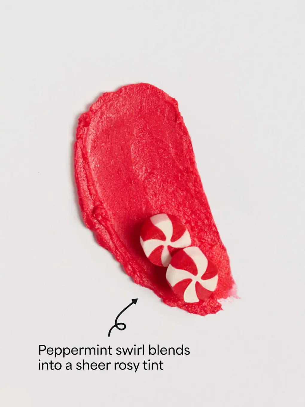 Sheer red balm swatch smeared on white with two red-and-white peppermint candies resting on it