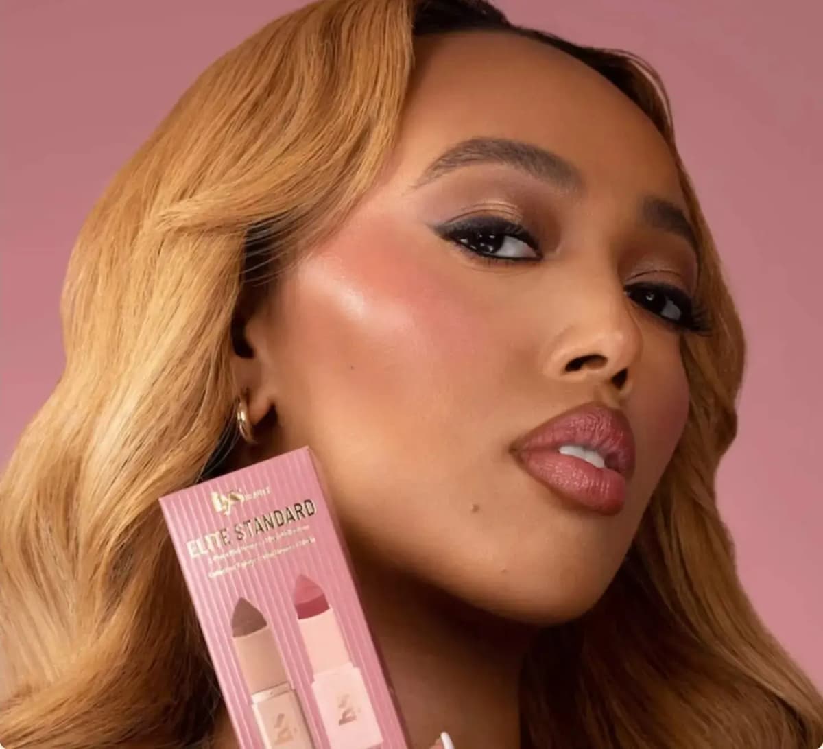 Model with glowing rosy cheeks and glossy mauve lips holds a pink Elite Standard bronze and blush box near her jaw