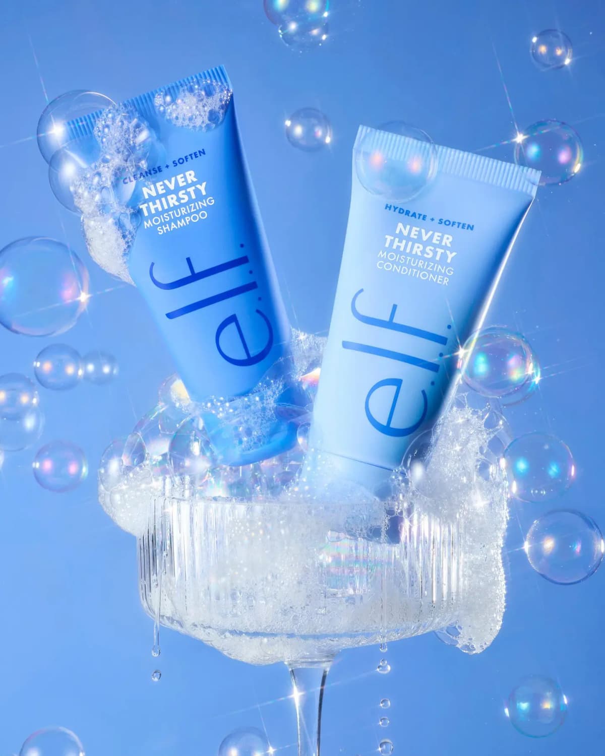 E.l.f. Never Thirsty mini shampoo and conditioner tubes in a foam-filled coupe glass amid iridescent bubbles on blue