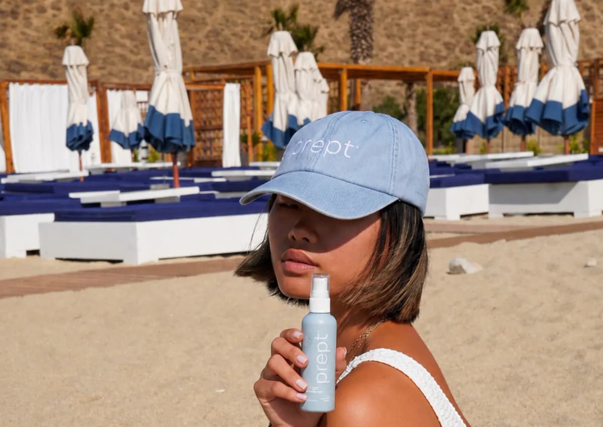 Woman in a light blue prept cap holds a prept go mist bottle on a sandy beach with blue-trimmed umbrellas behind