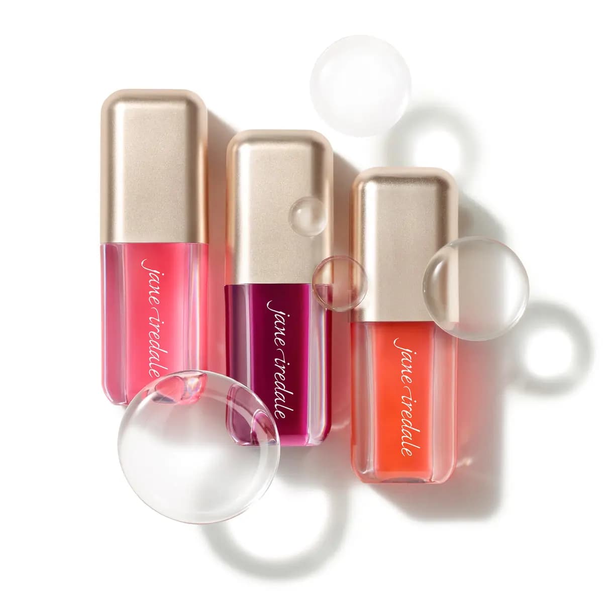 Three jane iredale lip oils in pink, berry and coral with gold caps, arranged among clear bubble-like spheres on white.