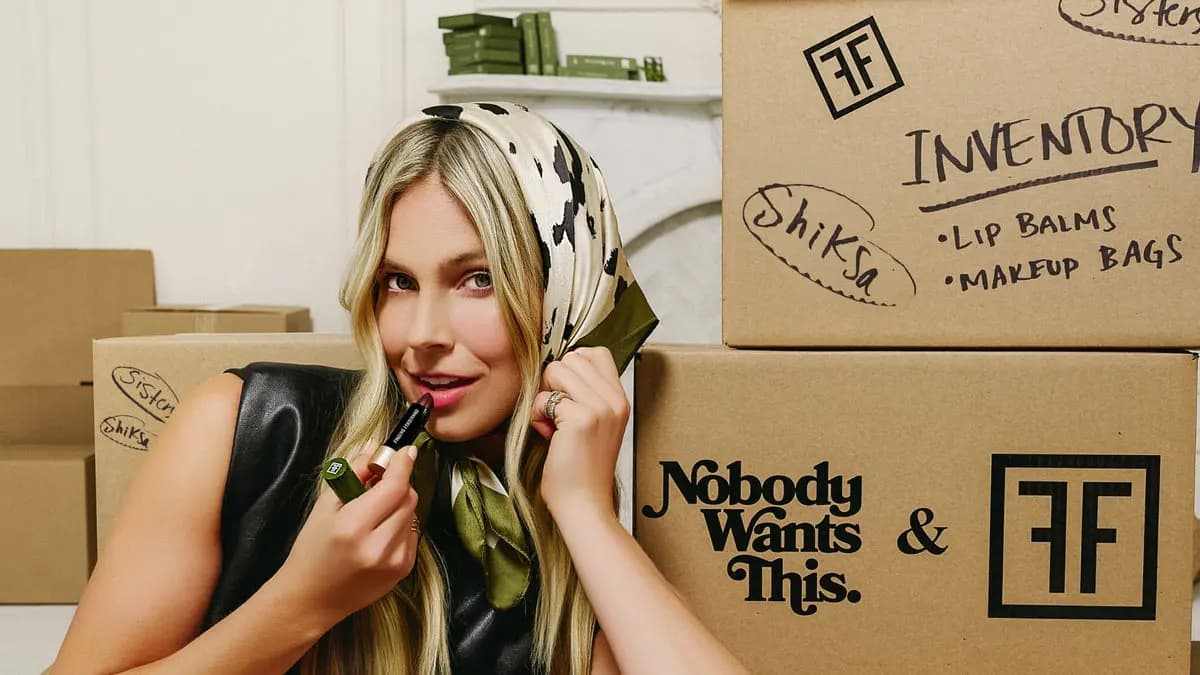 Blonde model in a cow-print headscarf holds the balm by cardboard boxes marked Inventory, Shiksa and Sisters