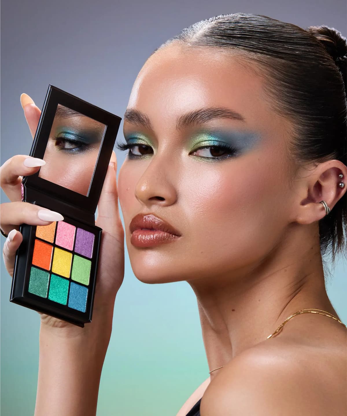 Model with green and teal shimmer eyeshadow holding the open palette, its mirror reflecting the model's eye makeup