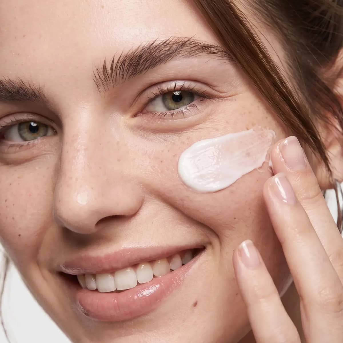 Smiling freckled model smoothing a swipe of white cream across the cheekbone with fingertips