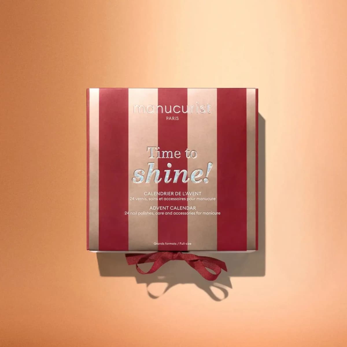 Overhead view of closed red and gold striped Time to Shine advent calendar box with red ribbon on peach background