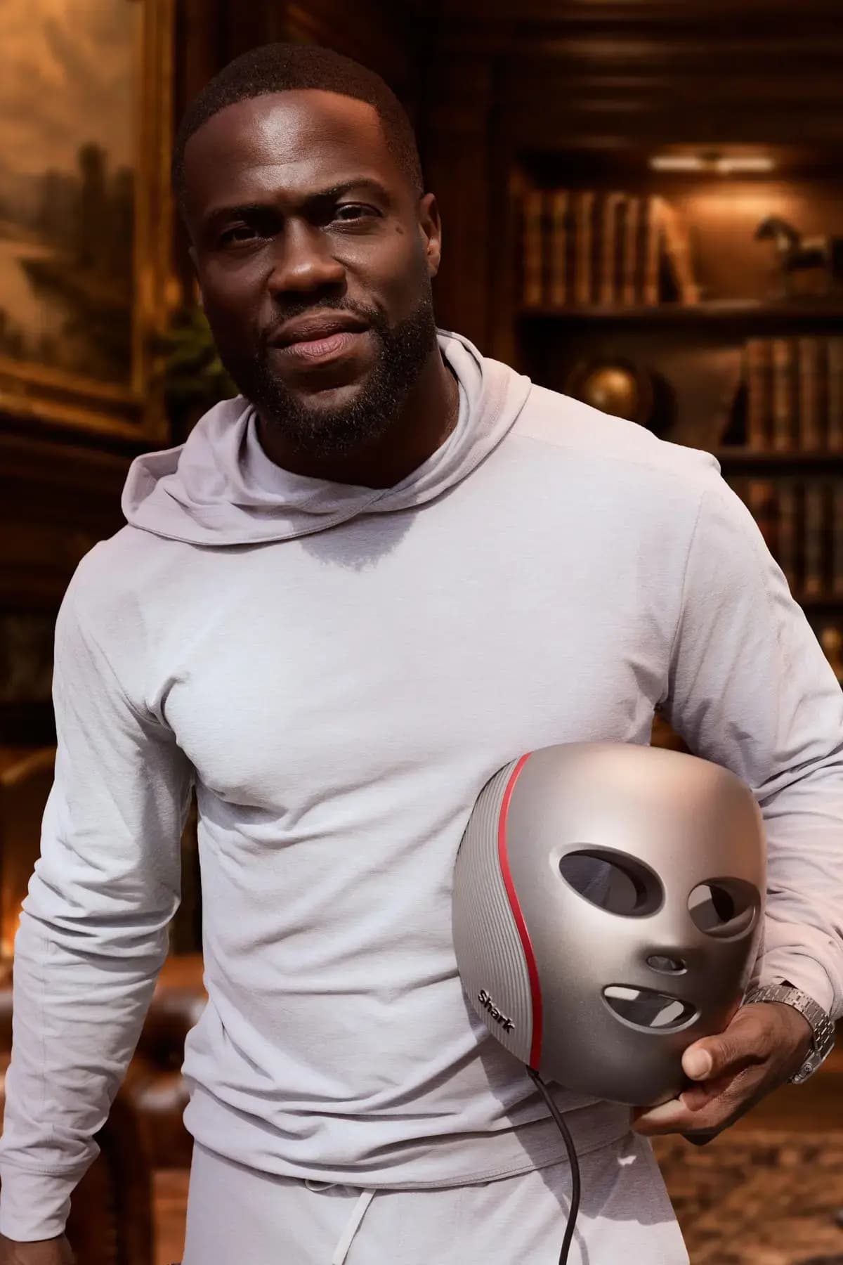Kevin Hart in a grey hoodie holds the gunmetal and red Shark CryoGlow mask under his arm in a wood-panelled library