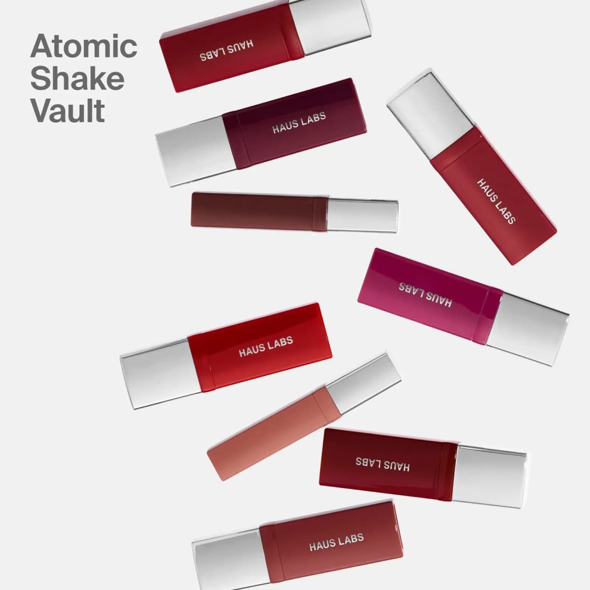 Nine Atomic Shake lacquer tubes in reds, berry and nude with silver caps scattered on a white background