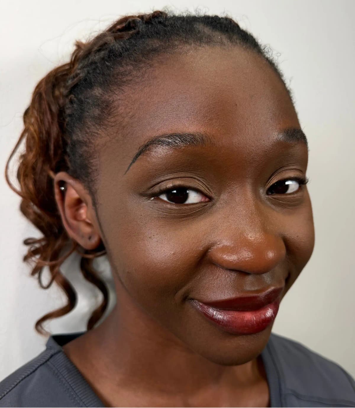Woman with deep skin tone smiling sideways, wearing a glossy deep red-brown lip with her hair pulled back in curls
