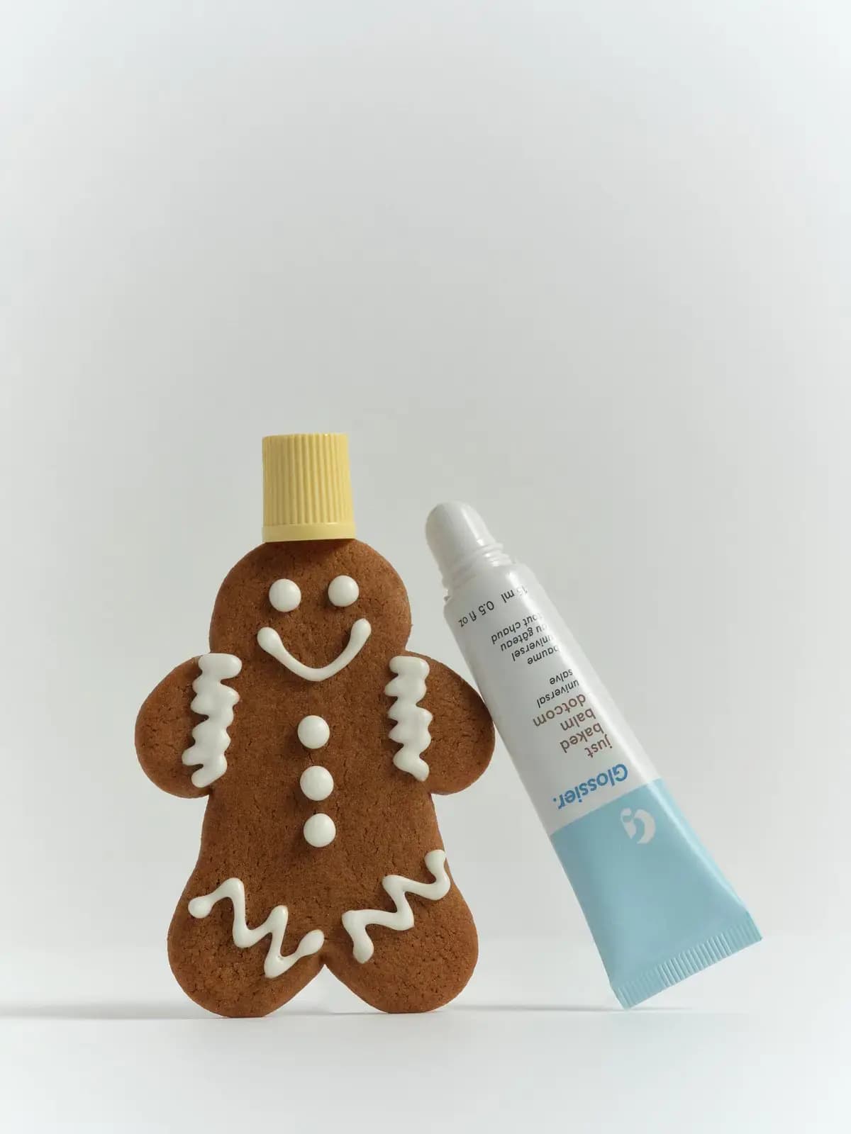 Glossier Just Baked Balm Dotcom universal salve tube leaning on an iced gingerbread man wearing its yellow cap as a hat